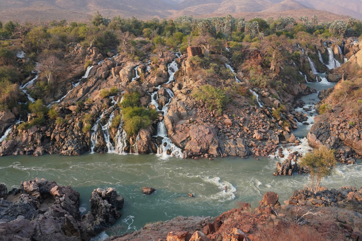 Landschaftsbild in der Vogelperspektive, das grün bewachsene braune Felsen zeigt, an denen kleine Wasserfälle in einen grünen Fluss fließen. 