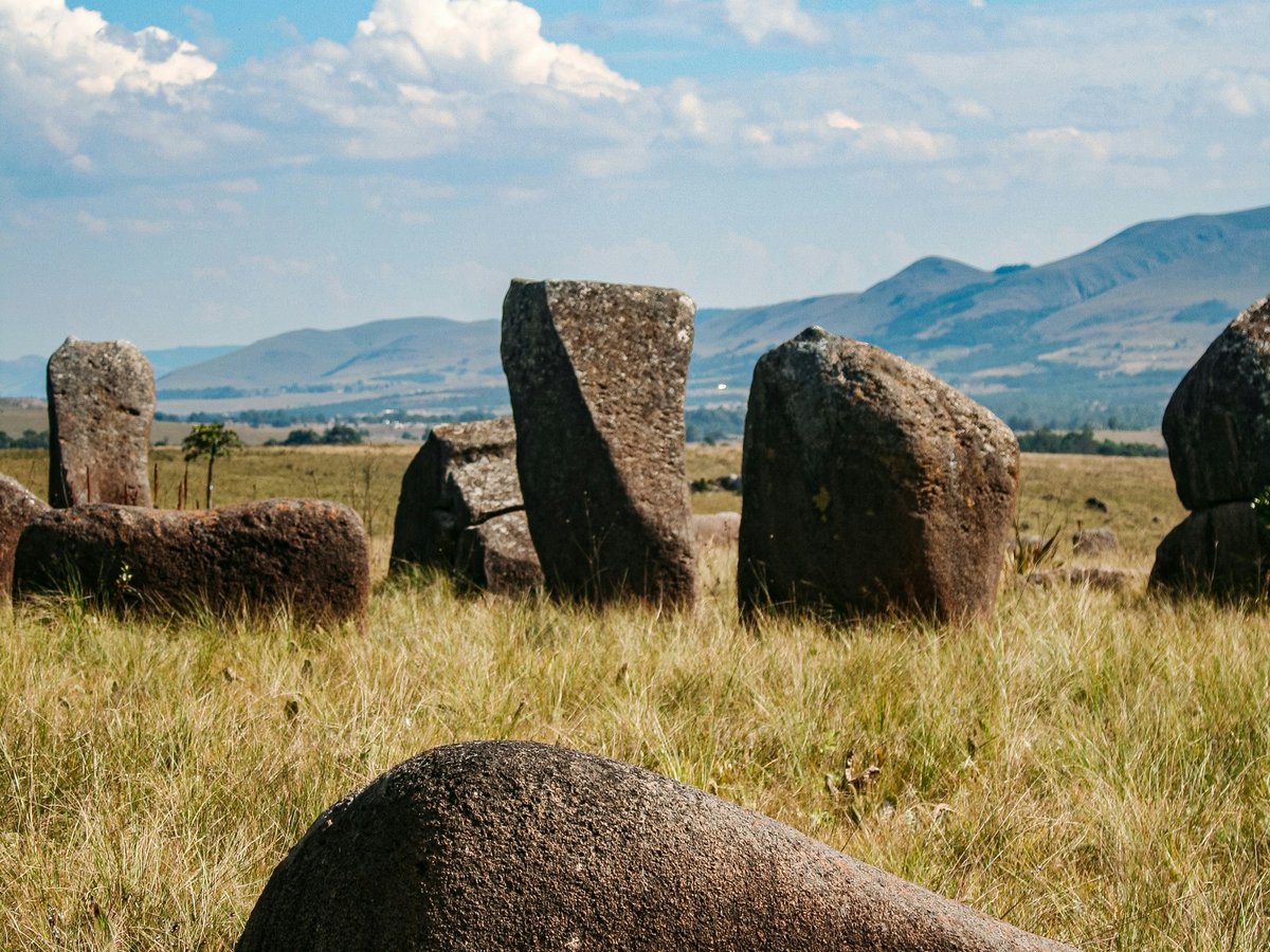 Gruppe großer, unregelmäßig geformter Steine auf einer Grasfläche vor Bergen unter blauem Himmel mit Wolken