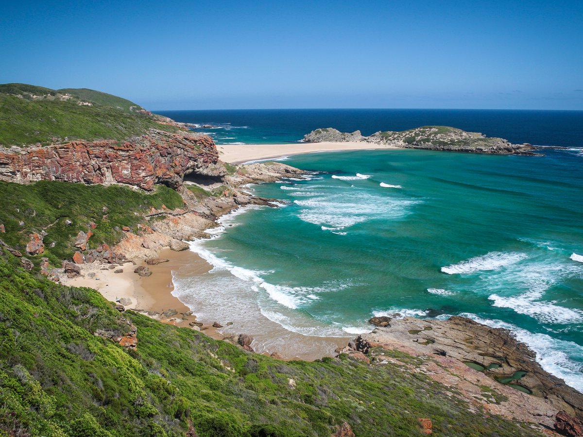 Küstenlandschaft mit felsigen Klippen, Sandstrand und türkisfarbenem Meer unter klarem Himmel.