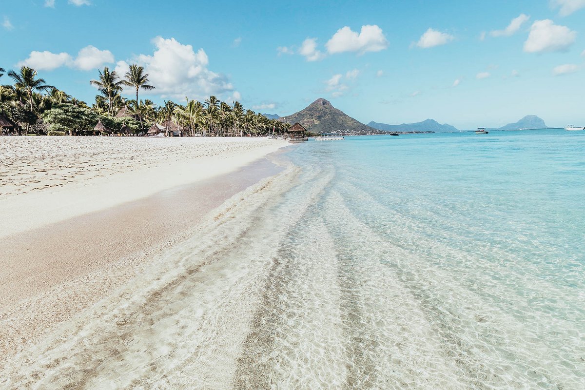 Kristallklares Wasser mit einem feinen Sandstrand am Rande eines dichten Palmenwaldes und im Hintergrund ein Berg.