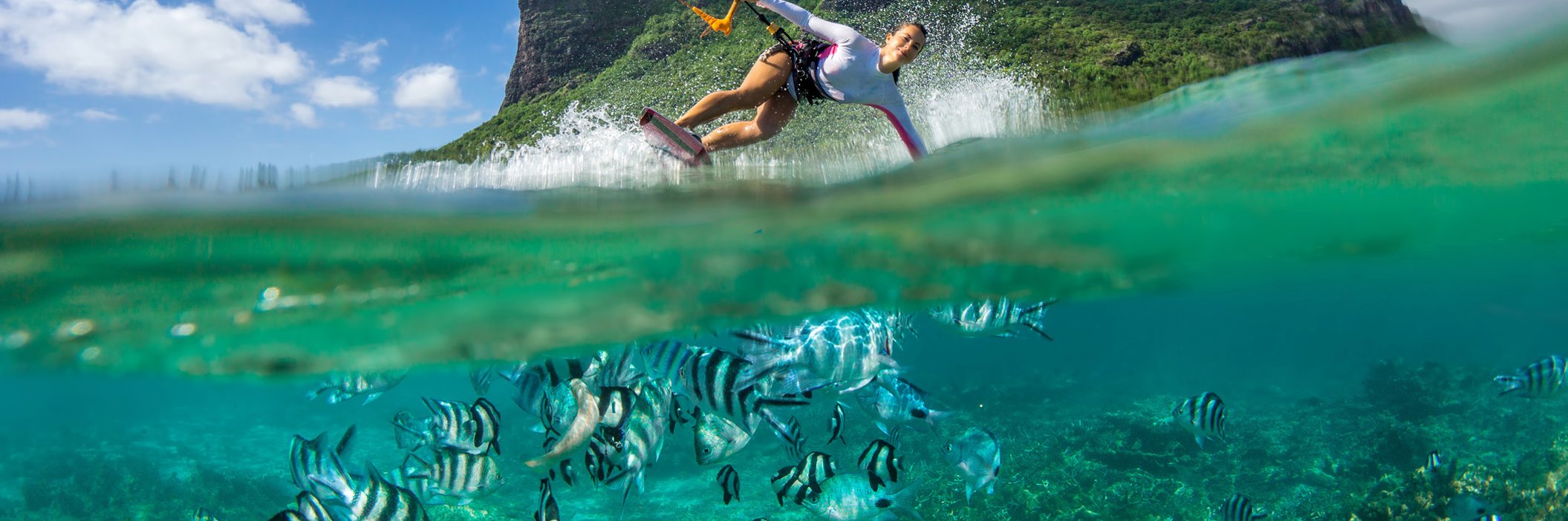 Dynamische Kitsurferin vor prägnantem Berg mit gleichzeitigem Blick auf Fischen unter Wasser. 