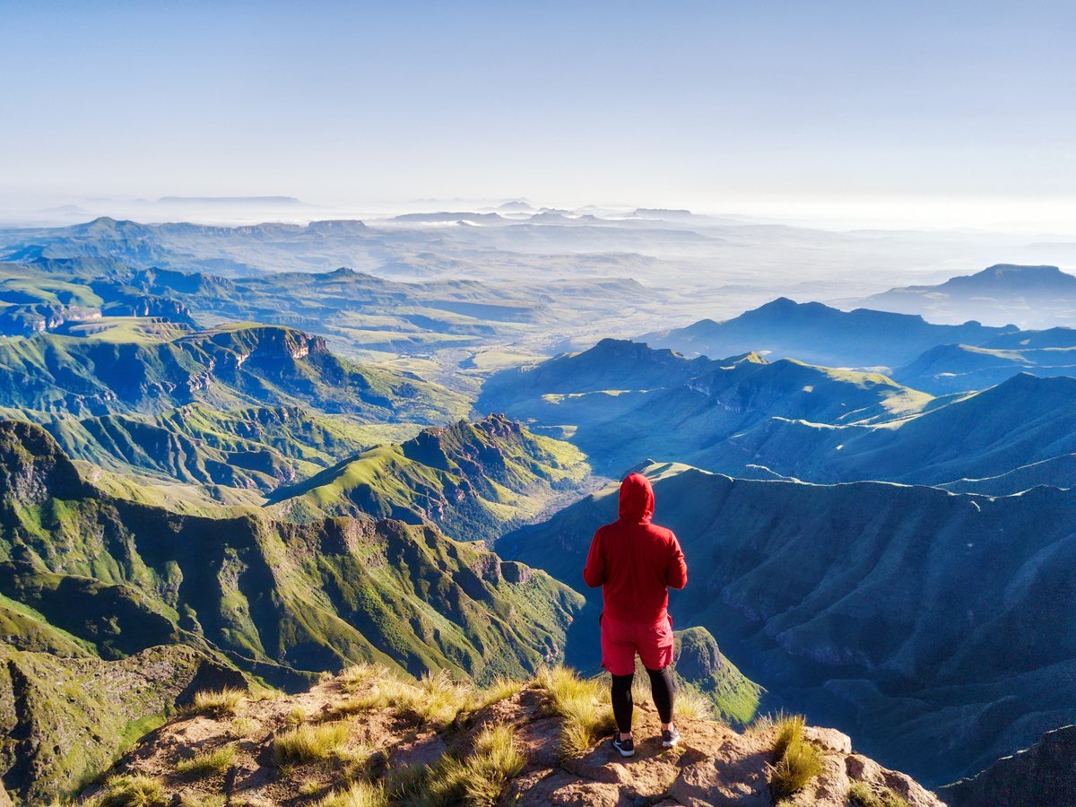 Person in roter Jacke steht auf einem Felsen und blickt auf eine weite Gebirgslandschaft
