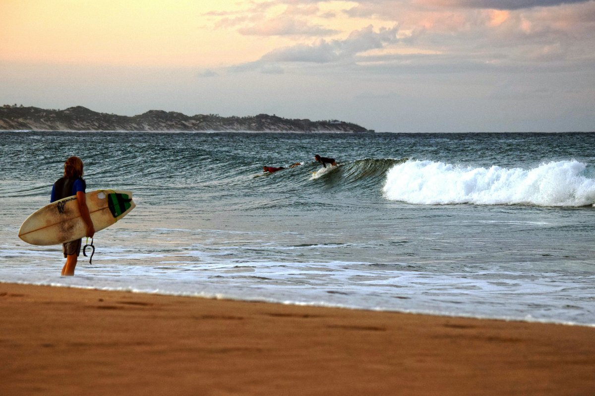 Surfer mit Surfbrett am Strand blickt auf das Meer mit Wellen und weiteren Surfern im Wasser