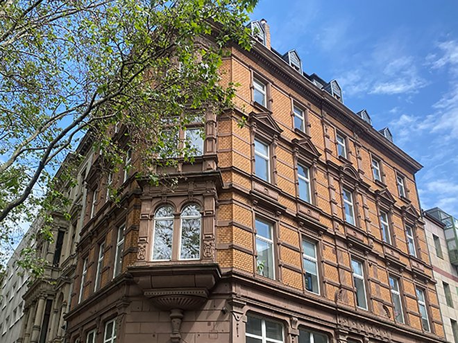 Mehrstöckiges Eckhaus mit großen Fenstern und verzierter Fassade, Baumäste ragen ins Bild.