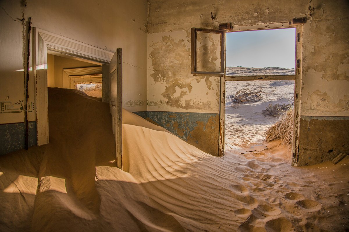 Das innere eines verlassenen Hauses, welches mit Sandbergen gefüllt ist und einen Blick auf Wüste frei gibt. 